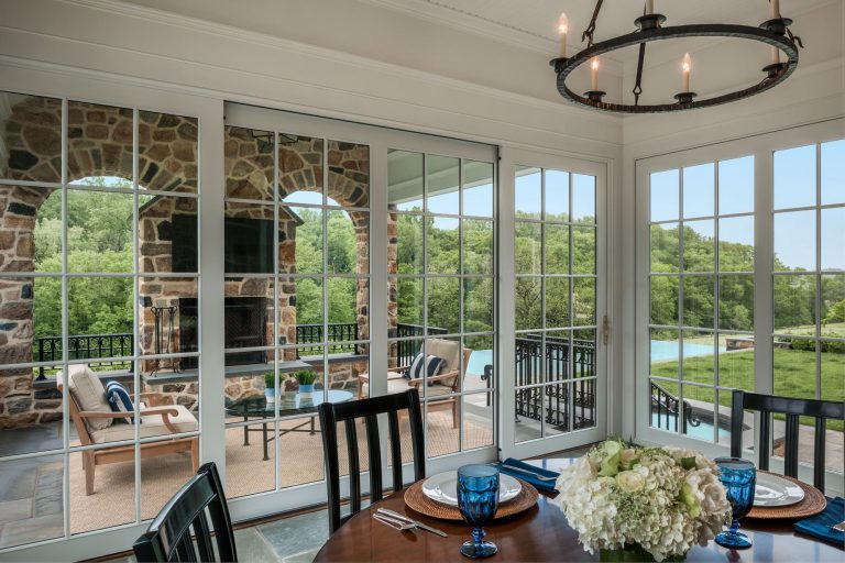 A bright dining area with a round wooden table set for four, featuring blue glassware and a white floral centerpiece. Large glass doors open to a stone patio with outdoor seating, overlooking green trees and a pool in the background.